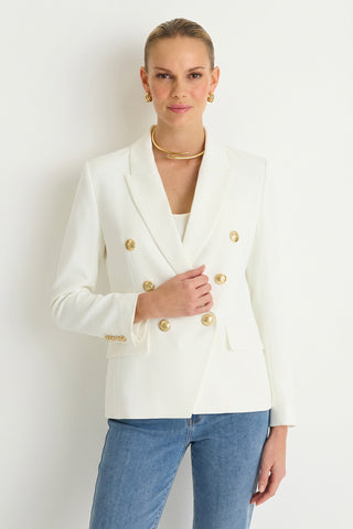 A woman wears the Sasha Double Breasted Blazer - White with gold hoop earrings, a gold necklace, blue jeans, and her hair pulled back, posing against a plain white background for a chic suiting look.