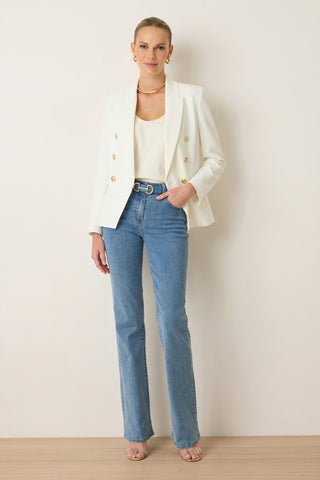 A woman stands against a plain wall wearing the Sasha Double Breasted Blazer in white, paired with a cream top, belted blue jeans, and nude heels. She accessorizes with hoop earrings, a gold necklace, and has her hair pulled back.