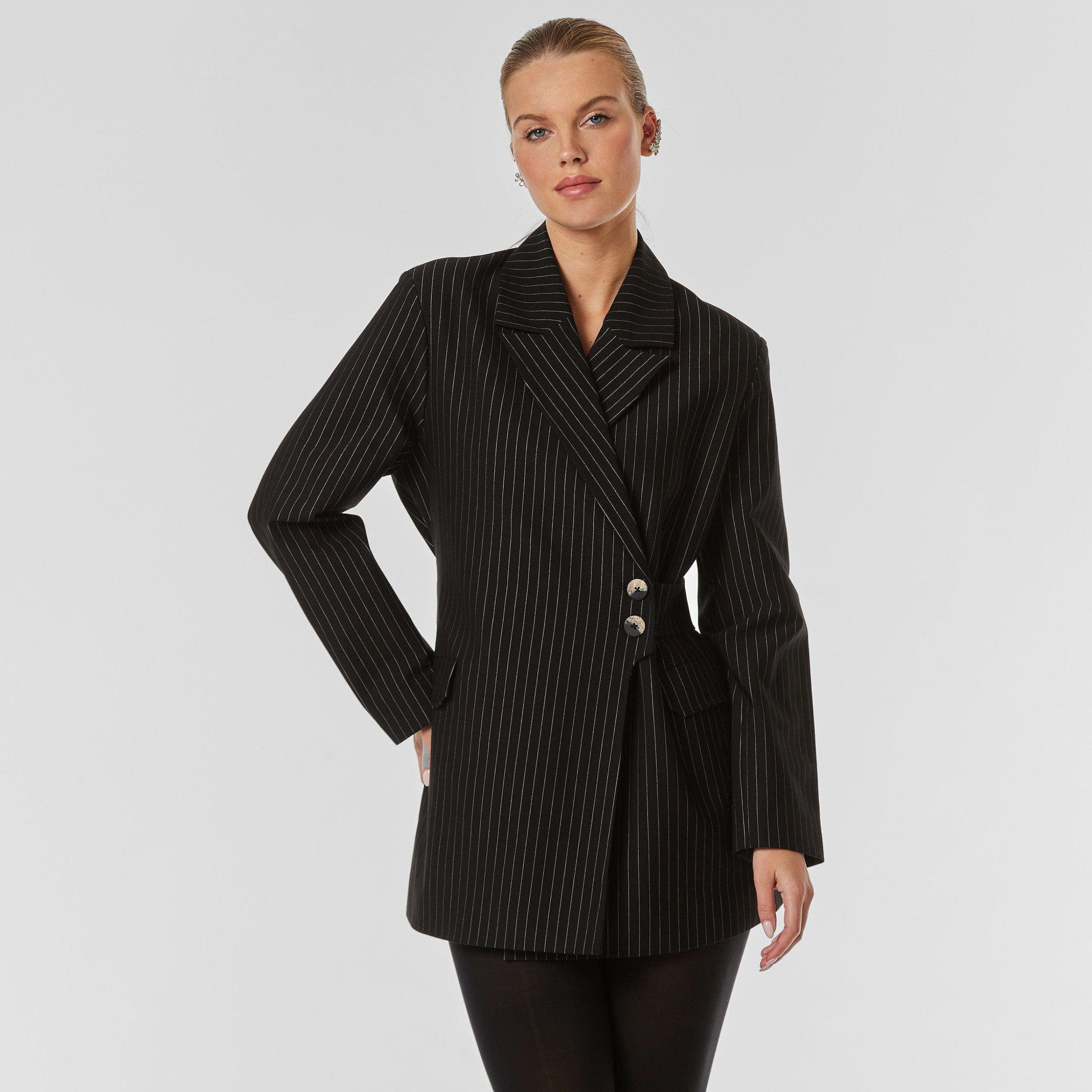 Front view of woman wearing black pinstripe blazer, featuring peaked lapel, flap pockets, detachable belt with button closure, flap pockets and silky lining.