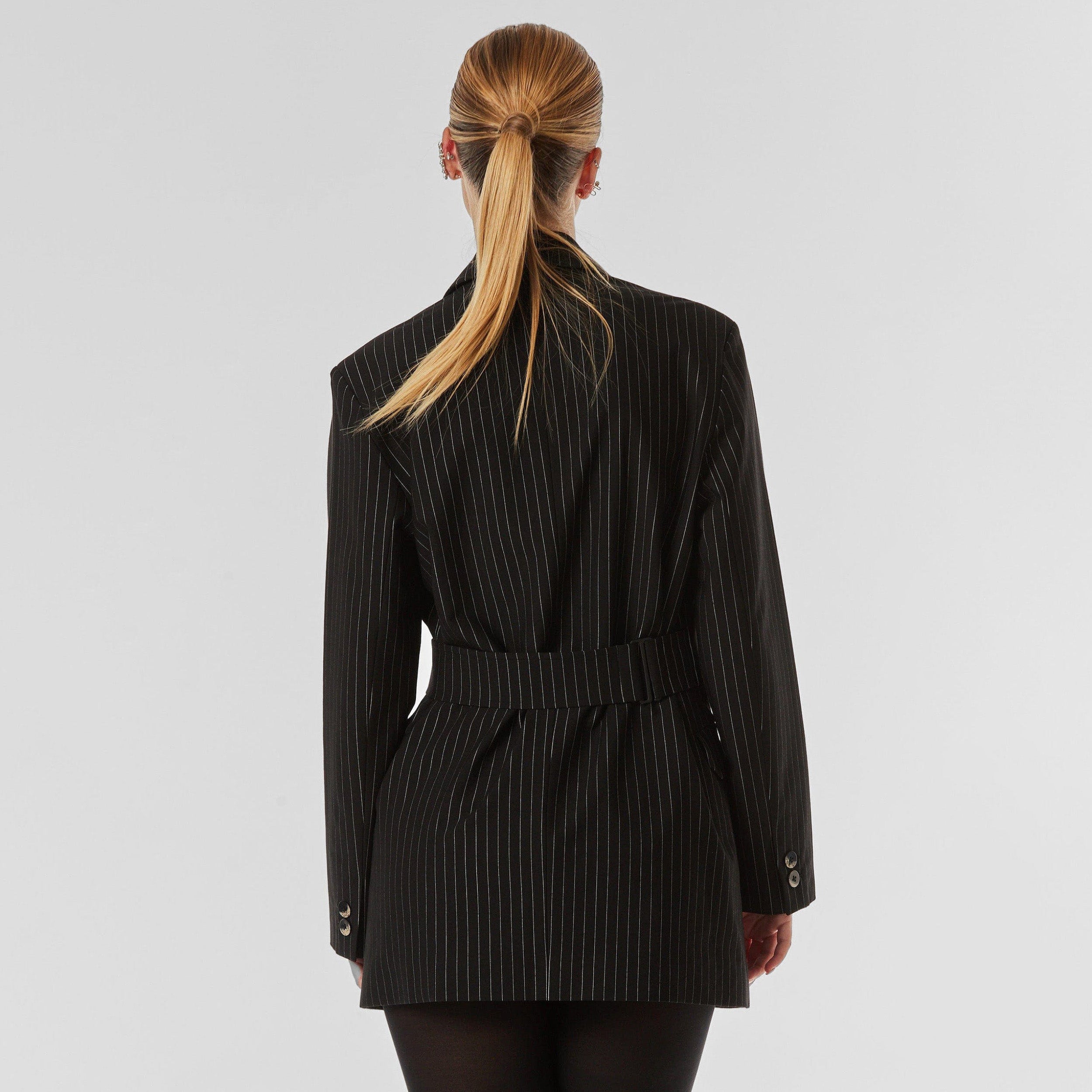 Rear view of woman wearing black pinstripe blazer, featuring peaked lapel, flap pockets, detachable belt with button closure, flap pockets and silky lining.