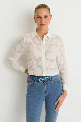 A woman stands against a white wall, wearing the Stelle Button Up - White—a sheer blouse adorned with a delicate leaf lace pattern that makes for an elegant, feminine layering piece. She pairs it with blue jeans, a belt featuring a gold buckle, and gold hoop earrings. Her hair is pulled back as she smiles softly.