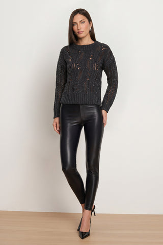 A woman stands against a plain wall wearing the Suzette Skinny Pant in black, paired with a black knit sweater and black heels, her arms relaxed by her sides.