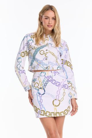 A woman faces forward wearing the Monaco Denim Skirt - Chain Print (Preorder), styled with a lightweight, long-sleeve cropped jacket featuring a colorful chain-link pattern on white. She has wavy blonde hair and a neutral expression.