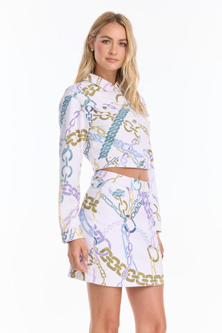 A woman stands against a plain white background wearing a lightweight, long-sleeve crop top and the Monaco Denim Skirt - Chain Print (Preorder), both featuring vibrant chain prints for a bold, statement look.