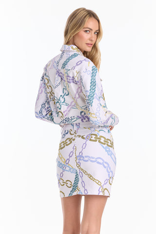 A woman with long blonde hair models the Monaco Denim Skirt - Chain Print (Preorder) as part of a matching set in blue, gold, and light purple chains. She stands facing away, looking over her shoulder against a plain white background.