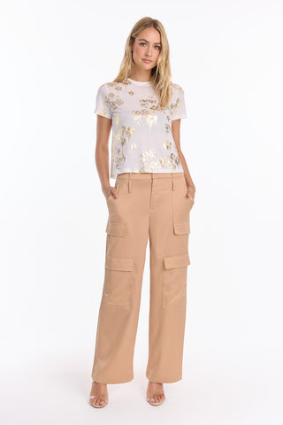 A woman with long blonde hair stands facing forward, wearing a white T-shirt and Milan Satin Cargo Pant - Gold (Preorder) wide-leg trousers with large pockets, hands in her pockets, against a plain white background.