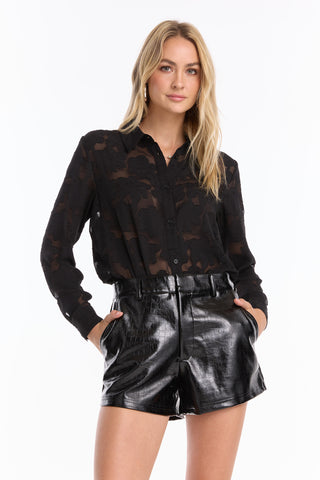 A woman stands against a white background, hands in her pockets, wearing a black sheer floral long-sleeve blouse and Jaclyn Leather Short - Black, looking at the camera.