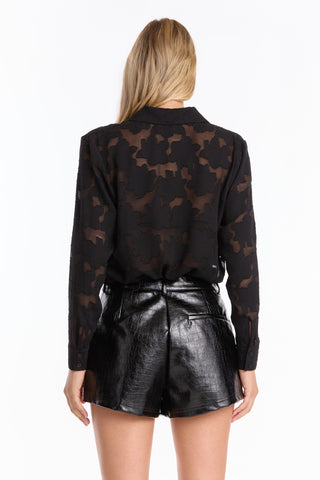 A woman with straight blonde hair stands facing away, wearing a sheer patterned long-sleeve blouse and the Jaclyn Leather Short - Black, featuring a high-waisted faux croco embossed design that elongates her legs.
