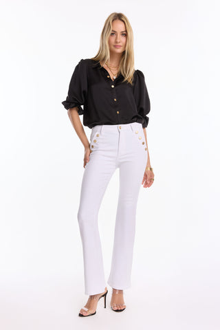 A woman stands confidently in the Monroe Silky Bodysuit - Black (Preorder), styled with rolled sleeves, high-waisted white pants with gold buttons, and open-toed heels against a plain white background—a chic summer look.