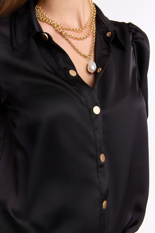 A person wearing the Monroe Silky Bodysuit - Black (Preorder), a luxe satin blouse with gold buttons, styled with layered gold chain necklaces featuring a large pearl pendant—a true summer dream look.