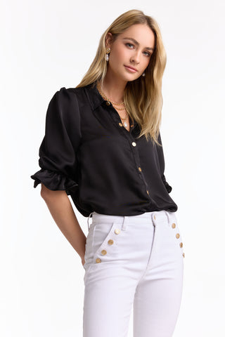 A woman with long blonde hair models the Monroe Silky Bodysuit in black (preorder) styled with white high-waisted pants with gold buttons, posing confidently against a plain white background and capturing effortless summer style.