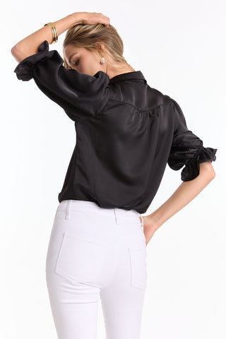 A woman faces away, wearing the Monroe Silky Bodysuit - Black (Preorder) in luxe satin with puffed sleeves and white high-waisted jeans. One hand rests on her head, the other on her hip, set against a plain white background.