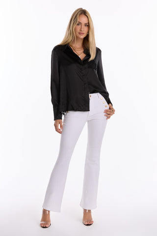 A woman stands confidently against a white background wearing the Amelia Silky Lace Blouse - Black (Preorder) with an open lace back, paired with white high-waisted flared pants, clear heels, layered necklaces, and long blonde hair.