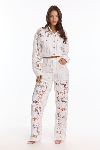 A woman faces forward in the Celeste Crochet Jean - White (Preorder), a pair of high-waisted, semi-sheer lace pants styled over a neutral underlayer, paired with open-toe heeled sandals.