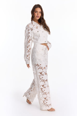 A woman with long brown hair and sandals models the Celeste Crochet Jacket - White (Preorder), paired with high-waisted wide-leg pants for a chic, semi-sheer lace look. She stands against a plain white background, showcasing a flattering silhouette.