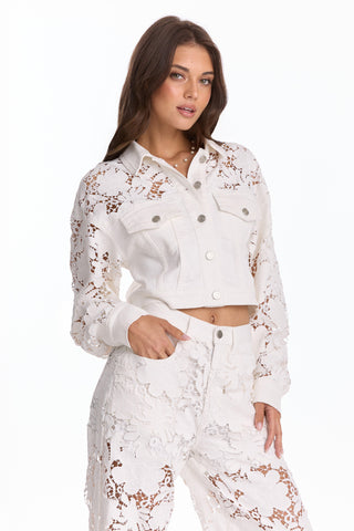 A woman confidently poses against a plain white background wearing the Celeste Crochet Jacket - White (Preorder) styled with high-waisted pants featuring lace and floral cutout details for a flattering silhouette.