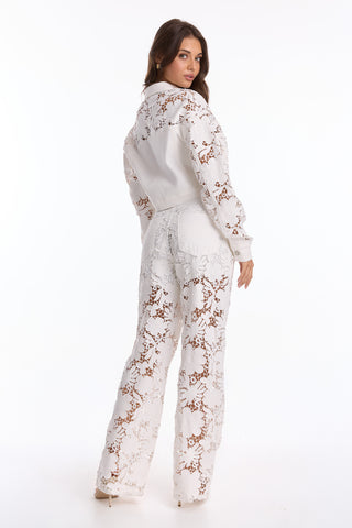 A woman poses against a plain white background, turned slightly to the side, wearing the Celeste Crochet Jean - White (Preorder), which features sheer floral lace panels and creates a flattering silhouette. She has long brown hair.