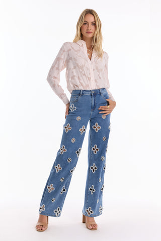 A woman stands confidently in the Stelle Button Up - Pearl, a light, semi-sheer floral lace blouse, paired with blue jeans featuring white and brown floral embroidery. She has long blonde hair and wears light heeled sandals.