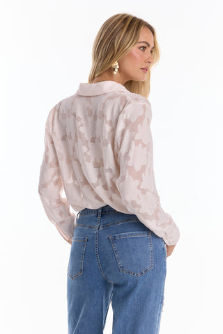 A woman with long blonde hair, wearing the Stelle Button Up - Pearl as a sheer leaf-patterned layering blouse, pearl drop earrings, and high-waisted blue jeans, stands facing away while looking over her shoulder.