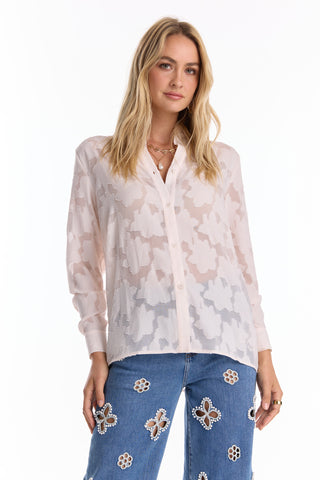 A woman wears the Stelle Button Up - Pearl, a sheer light pink floral lace blouse styled with blue jeans featuring large cut-out flower designs, standing against a white background—ideal for layering on any occasion.