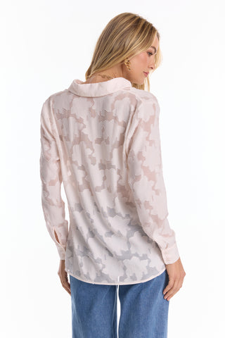A woman wears the Stelle Button Up - Pearl, a sheer, lightweight lace blouse in light pink with floral patterns and blue jeans, viewed from the back as she turns slightly to the side against a white background.