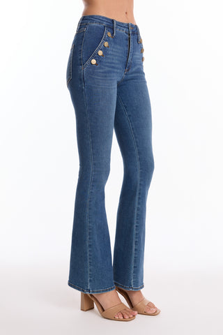Wearing the Allegra Bootcut Jean - Medium Blue (Preorder), high-waisted and flared with decorative front pocket buttons, paired with tan block-heeled sandals, the model stands against a plain white background for a vintage-inspired look.