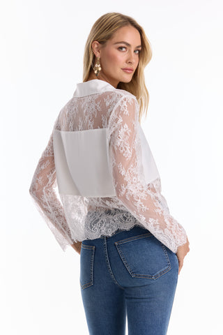A woman with long blond hair wears blue jeans and the Vienna Lace Blouse - White (Preorder), featuring a sheer lace front, solid back, and scalloped hem. She glances over her shoulder, showing off her dangling earrings.