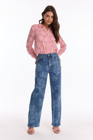 A woman faces forward in a light pink Stelle Button Up - Rose and high-waisted blue floral print jeans. She has long, wavy brown hair, wears open-toed heels, and stands against a white background.