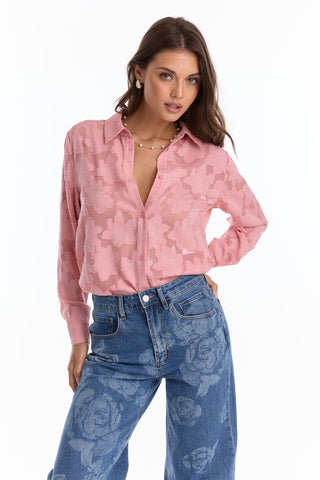 A woman with long brown hair and a neutral expression poses against a white background wearing the Stelle Button Up - Rose, a light pink floral blouse, paired with high-waisted blue jeans with large floral prints.