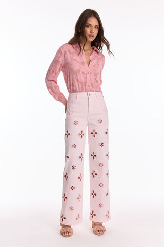 A woman stands against a white background wearing a light pink blouse with a floral pattern, blush Petite Kaia Pearl High Rise Jeans, and pink heeled sandals.