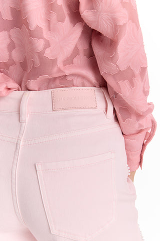 A person wears Petite Kaia Pearl High Rise Jean - Blush (Preorder), a light pink petite jean with a rectangular "THE NOLI SHOP" patch above the back pocket. Waist and upper legs are shown, paired with a textured pink floral blouse.