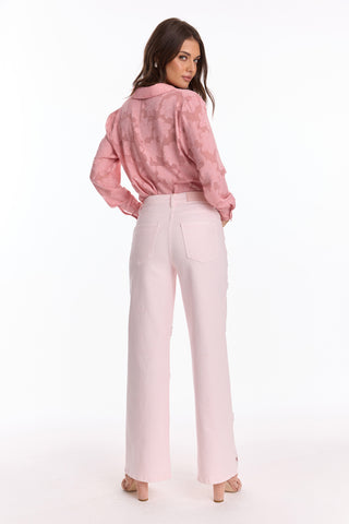 A woman stands facing away, looking over her shoulder. She wears the Petite Kaia Pearl High Rise Jean - Blush (Preorder) and a long-sleeve pink blouse with embroidered beaded flowers against a white background.