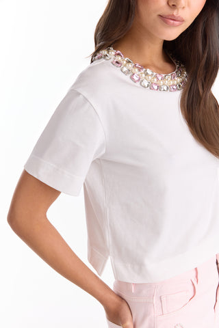 A woman wears the Crystal Naya T-Shirt - White, featuring a sparkling embellished neckline with large jewels. Paired with light pink pants, this chic cropped tee is perfect for a stylish summer look.