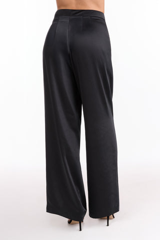 A person wearing the Valentina Satin Pant - Black, featuring a high waist and wide-leg silhouette, paired with black high heels, is shown from behind against a plain white background.