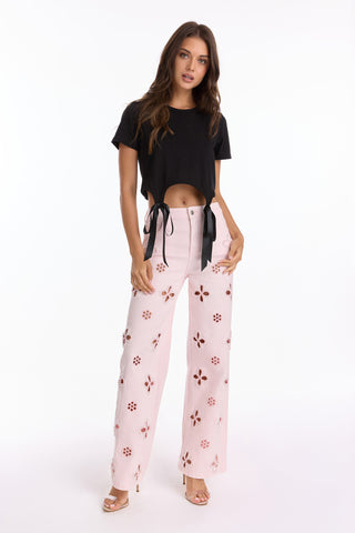 A woman with long brown hair stands against a white background wearing a black Diana Satin Bow Shirt and high-waisted, light pink Rosalie Jean pants with floral cut-out patterns, paired with heeled sandals.