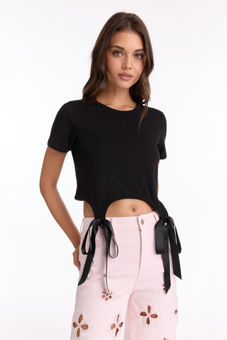 A woman wears the Diana Satin Bow Shirt - Black, a cropped cotton tee with satin bow ribbon ties at the waist, paired with light pink high-waisted pants with cut-out floral details, standing against a plain white background.