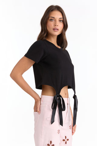 A woman wears the Diana Satin Bow Shirt - Black, a cropped cotton top with satin bow ties at the hem, paired with light pink high-waisted shorts with flower cut-outs, standing against a plain white background.