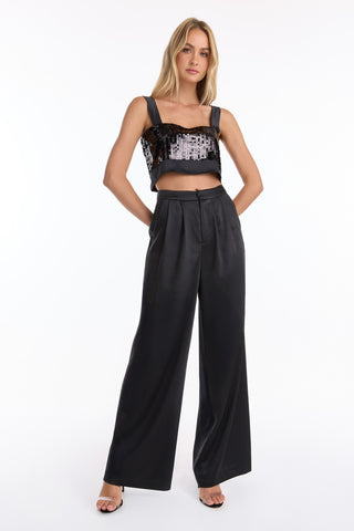 A woman with long blonde hair poses confidently against a white background, wearing a black sequin crop top with wide straps and the Valentina Satin Pant - Black, which features a high waist and a flattering wide-leg silhouette.