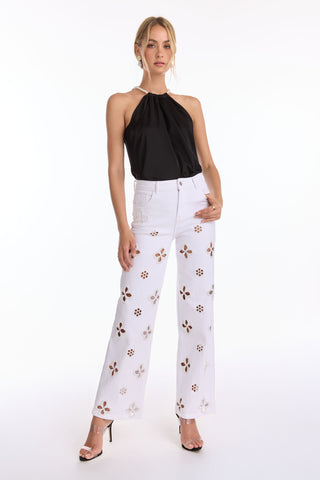 A woman poses in the Mika Silky Halter Top - Black (Preorder) with high-waisted white wide-leg pants featuring brown floral cutouts and clear high heels, embracing bold style against a plain white background.