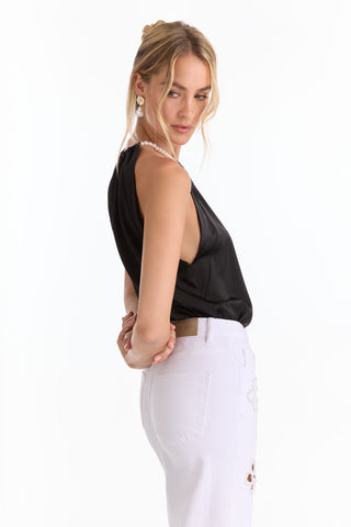 A woman with blonde hair in a loose updo wears the Mika Silky Halter Top - Black (Preorder) with white high-waisted pants, pearl earrings, and a pearl necklace, turning over her shoulder against a plain white background—statement style.