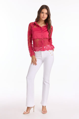 A woman with long brown hair and a neutral expression stands against a white background, wearing the Vienna Lace Blouse - Fuchsia (Preorder), featuring sheer long sleeves and a scalloped hem, paired with white buttoned pants and nude heels.