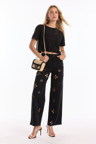 A woman stands confidently in a black cropped T-shirt, Kaia Pearl High Rise Jean - Black with embroidered beaded flowers, clear high-heeled sandals, and a small woven crossbody bag with a chain strap.