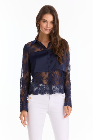A woman wears the Vienna Lace Blouse - Navy (Preorder), a sheer, elegant long-sleeve top with a solid chest panel, paired with white high-waisted pants with gold buttons, standing against a plain white background.