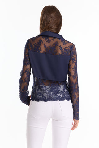 A woman faces away wearing white pants and the Vienna Lace Blouse - Navy (Preorder), a chic top featuring sheer lace sleeves, elegant back detailing, and a solid navy shoulder panel for a refined look.