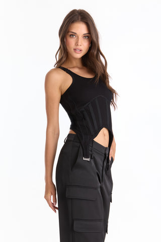 A woman with long brown hair poses confidently against a white background, wearing the fitted black Kiara Garter Tank, showcasing its corset-inspired details with one hand by her side, paired with high-waisted black cargo pants.