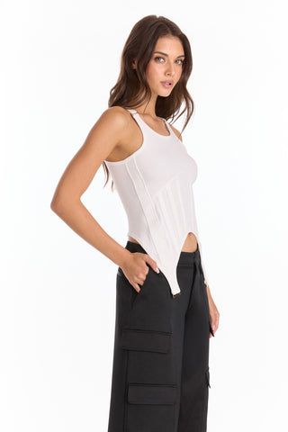 A woman with long brown hair stands against a plain white background, wearing black cargo pants and the Kiara Garter Tank - White, crafted from rib fabric with a corset-inspired design, her hands in her pockets.
