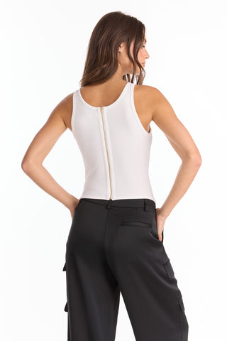 A woman faces away in a white Kiara Garter Tank with ribbed texture and back zipper, paired with black cargo pants. Her hair is down, arms at her sides, set against a plain white background.