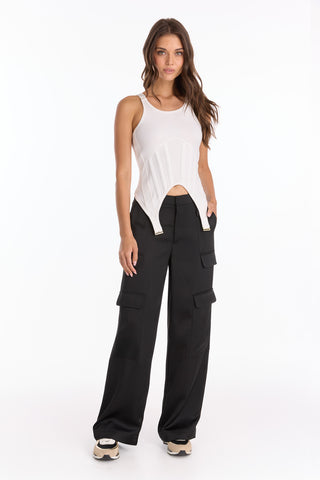 A woman with long wavy brown hair and a neutral expression wears the Kiara Garter Tank - White, styled with black cargo pants featuring large pockets and beige sneakers, against a white background.