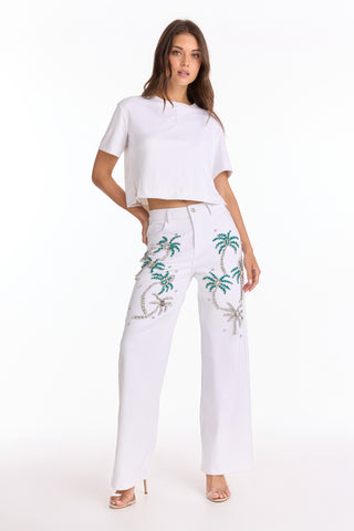A woman stands confidently in a white short-sleeve crop top and Crystal Palm High Rise Jean - White, featuring a blue and silver palm pattern. She poses with one hand on her hip, wearing open-toed heels and 4-way stretch denim.