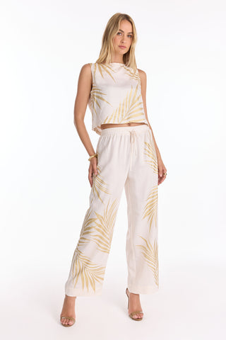 A woman stands confidently in the Sola Linen Pant - Gold Palm (Preorder), featuring a light beige palm leaf print and wide-leg silhouette. Ships June 25—pre-order now. She pairs it with a sleeveless top, open-toe heels, and long blonde hair against a white background.